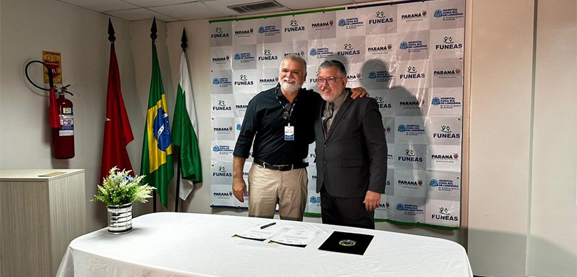 Fotografia de dois homens posando sorrindo abraçados em frente a um banner com os logos do Gover...