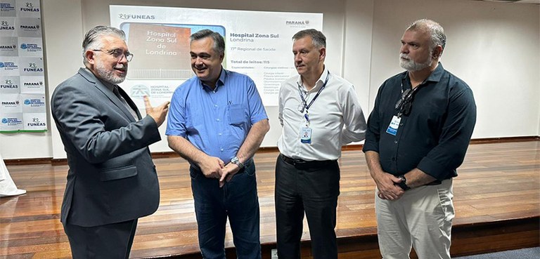 Fotografia de quatro homens conversando. Todos vestem roupas formais. Ao fundo, um telão, uma pa...