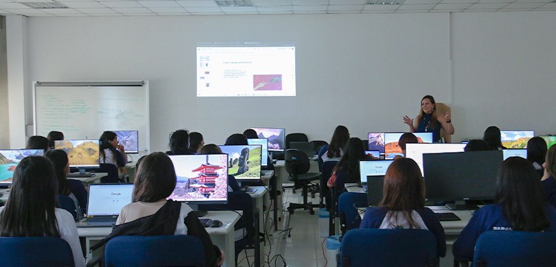 Fotografia de uma mulher em pé falando diante de diversas estudantes, que aparecem sentadas em c...