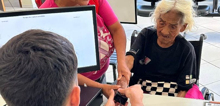 Fotografia de uma mulher idosa e indígena em uma cadeira de rodas coletando a biometria durante ...