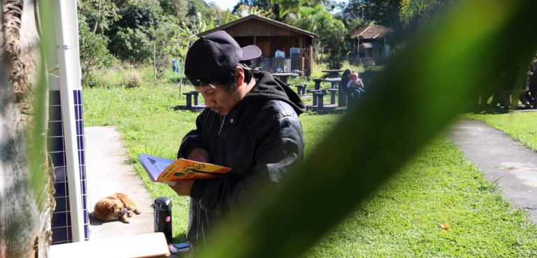 Fotografia de um homem indígena lendo a cartilha do TRE-PR. Ele usa boné e casaco pretos. Ao fun...