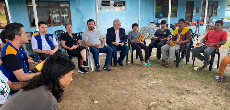 Fotografia de diversas pessoas, entre homens e mulheres, sentadas em roda. Ao fundo, há um edifí...