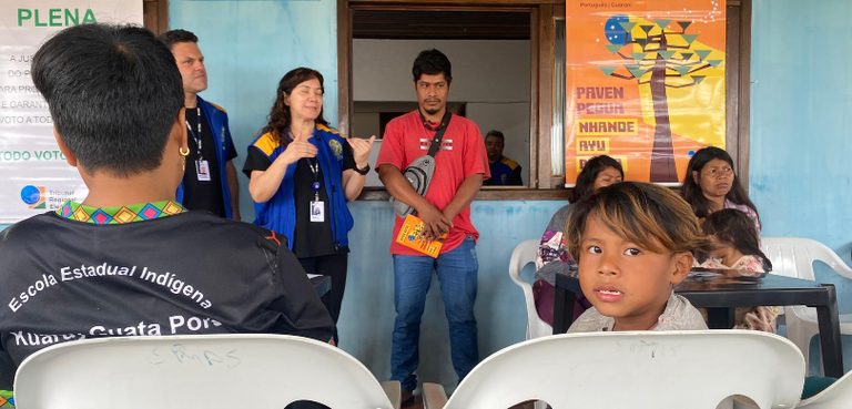 Fotografia de três pessoas em pé diante de outras pessoas sentadas em cadeiras brancas ao redor ...