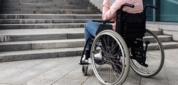 Fotografia de uma pessoa que usa cadeira de rodas parada de frente para uma escada. Não é possív...