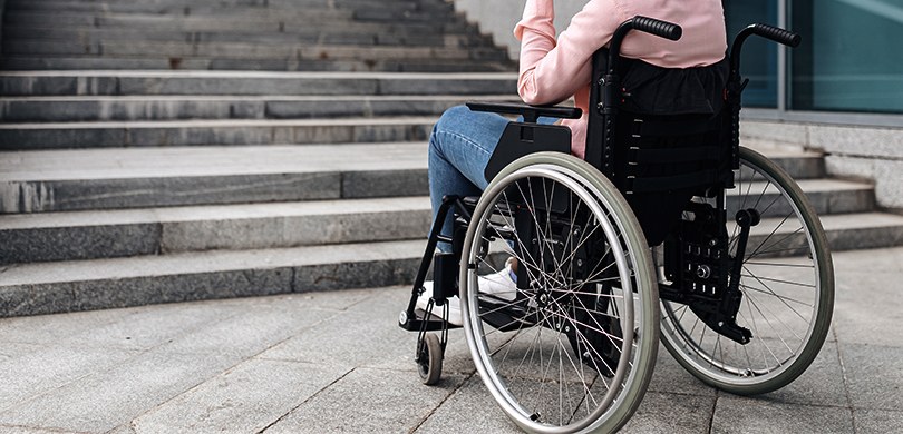 Fotografia de uma pessoa que usa cadeira de rodas parada de frente para uma escada. Não é possív...