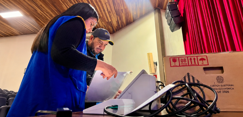 Fotografia na qual se vê uma mulher e um homem manuseando urnas eletrônicas sobre uma mesa. Em p...