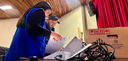 Fotografia na qual se vê uma mulher e um homem manuseando urnas eletrônicas sobre uma mesa. Em p...