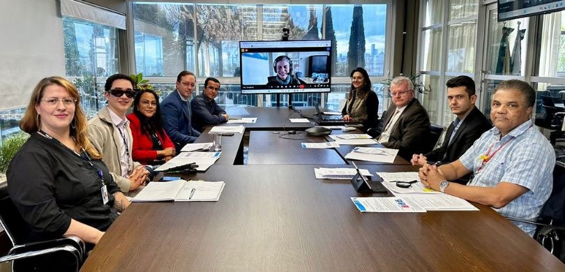Fotografia em que são vistas nove pessoas em torno de uma mesa de madeira posando. Ao fundo, obs...
