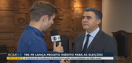 Fotografia de um senhor de terno e gravata dando entrevista para um repórter de televisão