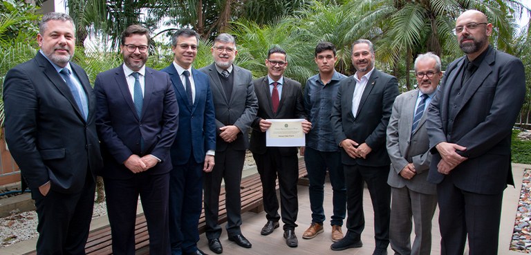 Fotografia de nove homens posando sorrindo. Todos usam roupas formais. O homem ao centro segura ...