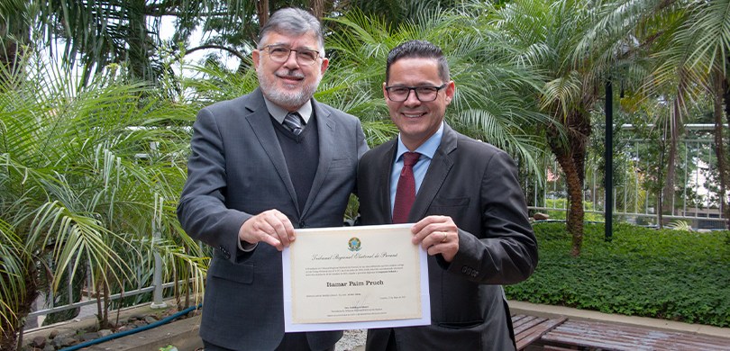 Fotografia de dois homens de terno posando para foto sorrindo enquanto seguram um diploma. Ao fu...