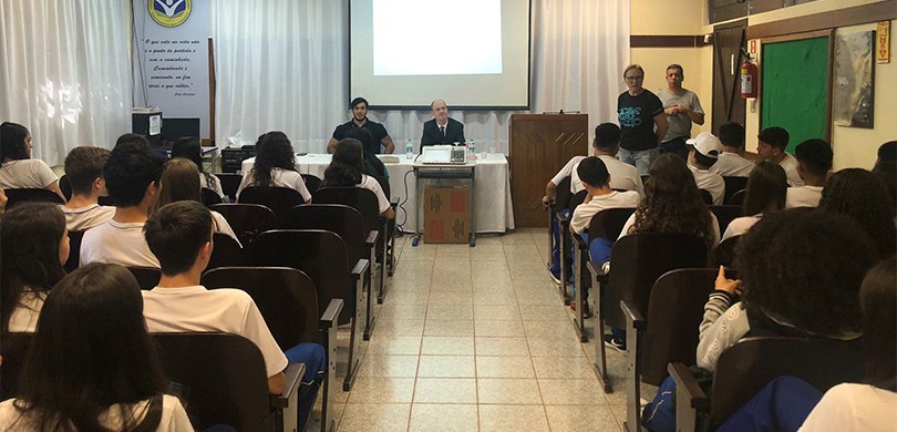 Em uma sala de aula, dois homens palestram para estudantes uniformizados com camiseta branca e c...