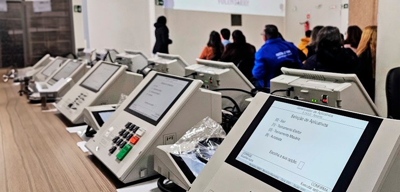 Fotografia de várias urnas eletrônicas sobre uma mesa de madeira. Ao fundo, desfocado,há uma mul...