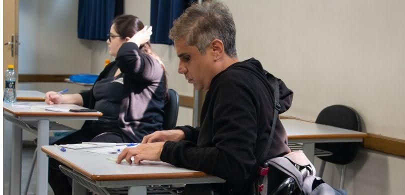 Fotografia em que se observam duas pessoas sentadas em carteiras escolares individuais dentro de...