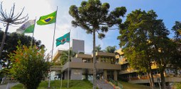 Fotografia do edifício-sede do TRE-PR, atrás das bandeiras do Paraná, Brasil e Curitiba e de uma...