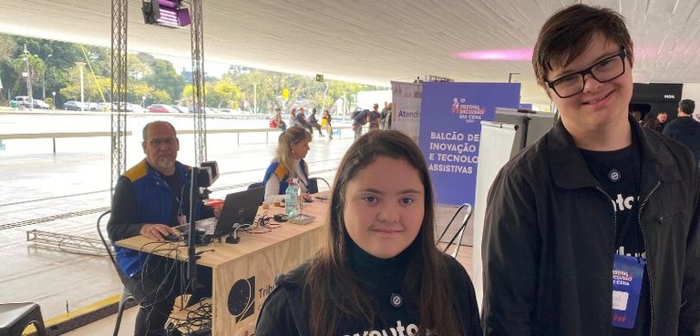 Fotografia que destaca, em primeiro plano, à direita, uma moça e um rapaz com síndrome de Down. ...