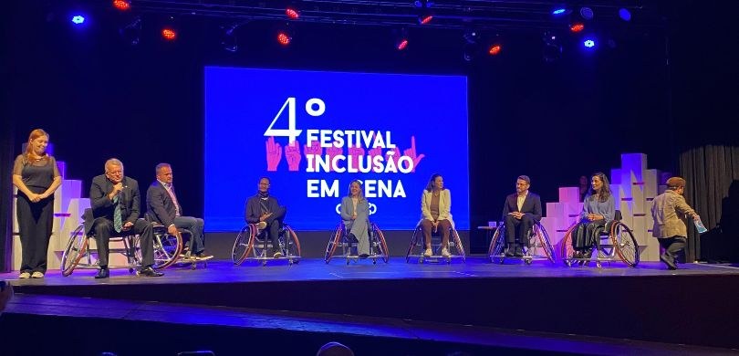 Fotografia em que aparecem pessoas sentadas em cadeiras de rodas em cima de um palco. Ao fundo, ...