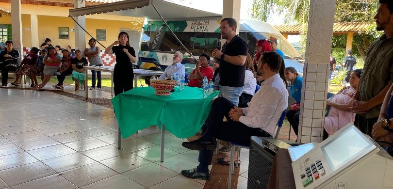 Fotografia em que se observa um grupo de aproximadamente vinte pessoas reunidas em um espaço cob...