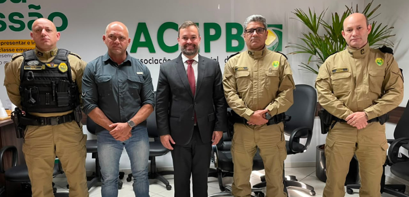 Fotografia em que se observam cinco homens alinhados lado a lado em um ambiente interno. Três de...