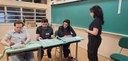 Fotografia de uma seção eleitoral. Há três mesários sentados atrás de mesas escolares e uma elei...