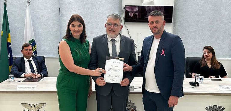 Fotografia de uma mulher e dois homens posando sorrindo segurando juntos um documento. Ao fundo,...
