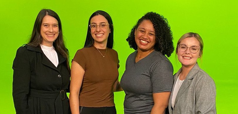 A foto mostra quatro mulheres sorrindo para a câmera. Elas estão abraçadas, uma do lado da outra...