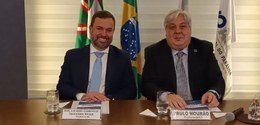 Fotografia na qual se observam dois homens sentados lado a lado atrás de uma mesa de madeira, po...