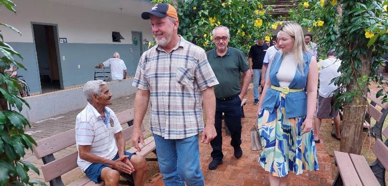Homens e mulheres passam por um corredor, em que há uma passarela de madeira com flores amarelas...