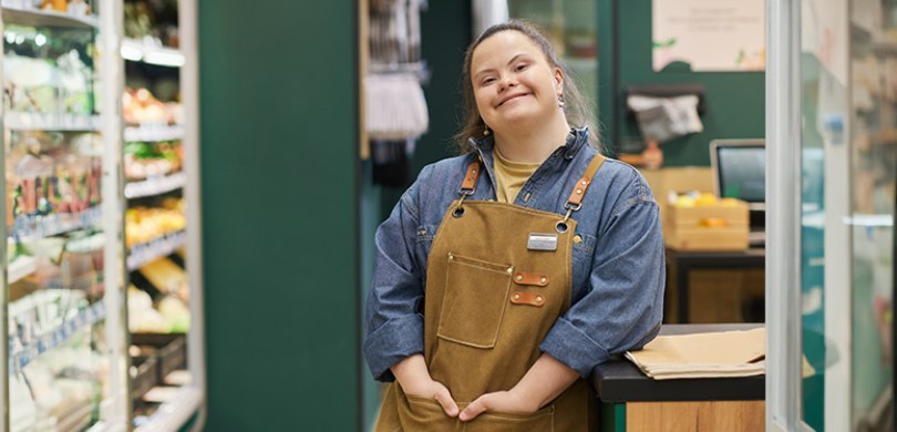 Em um supermercado com paredes verdes, uma mulher com síndrome de down sorri para a foto encosta...