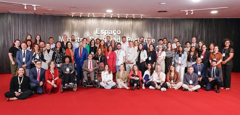 Fotografia de várias pessoas posando em pé sobre um tapete vermelho. Ao fundo, observa-se uma pa...