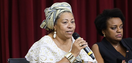 A fotografia mostra uma televisão e atrás um painel e uma cortina da cor vermelha. A tevê mostra...