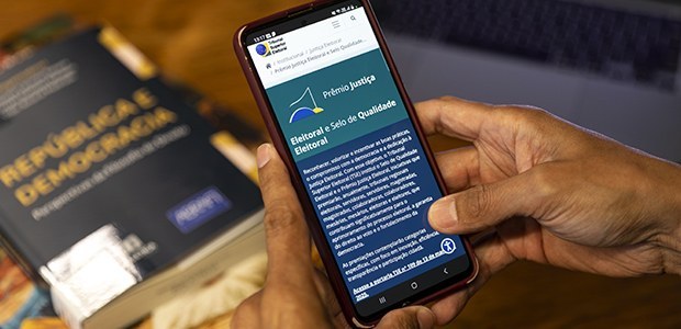 Fotografia das mãos de uma pessoa mexendo em um smartphone, que está aberto na página das premia...