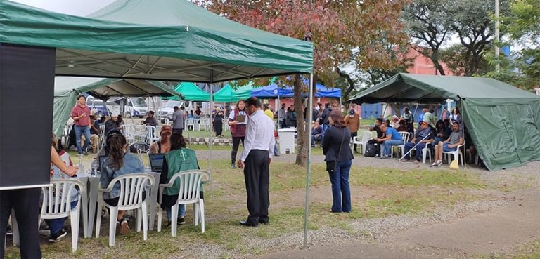 A fotografia mostra várias tendas dispostas em um espaço aberto com árvores em volta. Há bastant...