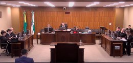 Fotografia em plenário presencial de tribunal, com tora e bancadas em madeira. Mais ao centro, h...