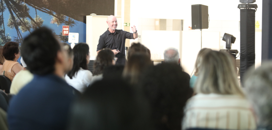 A Diretora-Geral Solange Maria Vieira fala para a plateia segurando um microfone. Atrás dela, um...