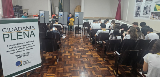 Fotografia de uma mulher em pé palestrando para jovens uniformizados em uma sala de aula com par...