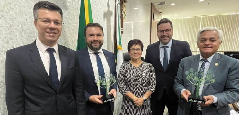 Fotografia de quatro homens de terno e uma mulher ao centro, que usa um vestido branco e preto. ...