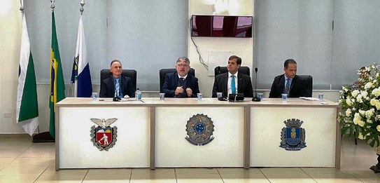Fotografia de quatro homens sentados atrás de uma mesa de autoridades. Todos usam terno. Ao redo...