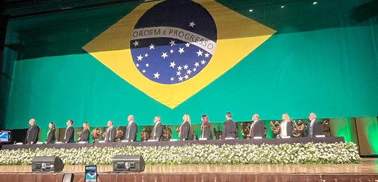 Fotografia da mesa de autoridades no palco de um auditório. Ao fundo, a bandeira do Brasil.