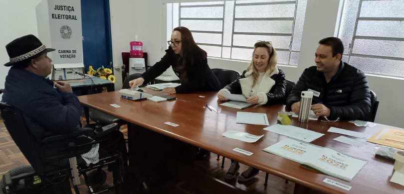 Fotografia em ambiente interno que retrata quatro pessoas em torno de uma mesa de madeira. À esq...