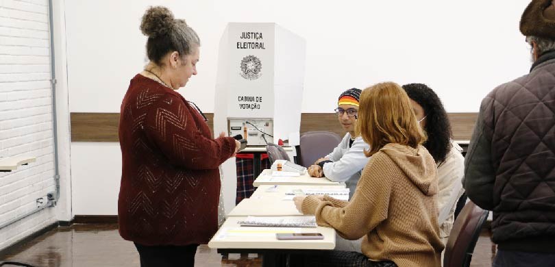 Fotografia de um homem e duas mulheres, de perfil, sentados em uma mesa em frente aos cadernos d...