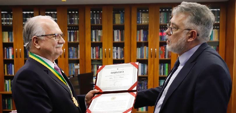 Fotografia de dois homens de terno se olhando, enquanto seguram juntos um diploma amarelo, com d...