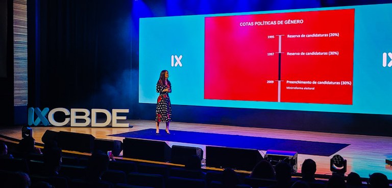 Fotografia de uma mulher diante de um telão em um auditório que se lê “Cotas Políticas de Gênero...