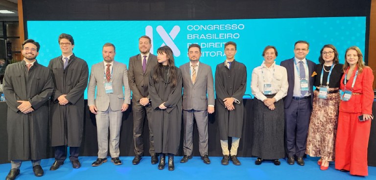 Fotografia de 11 pessoas, entre homens e mulheres, posando sorrindo em um palco. Ao fundo, há um...