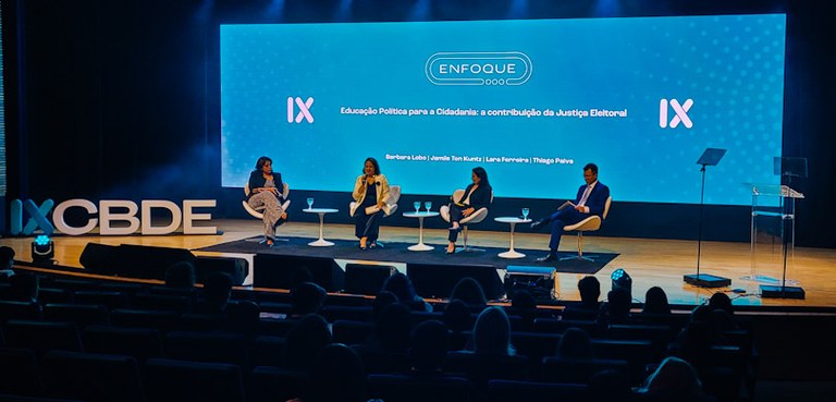 Fotografia de quatro pessoas sentadas em cadeiras em cadeiras brancas no palco de um auditório. ...