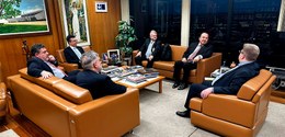 Fotografia de seis homens de terno sentados em poltronas marrons, que estão alinhadas em círculo...