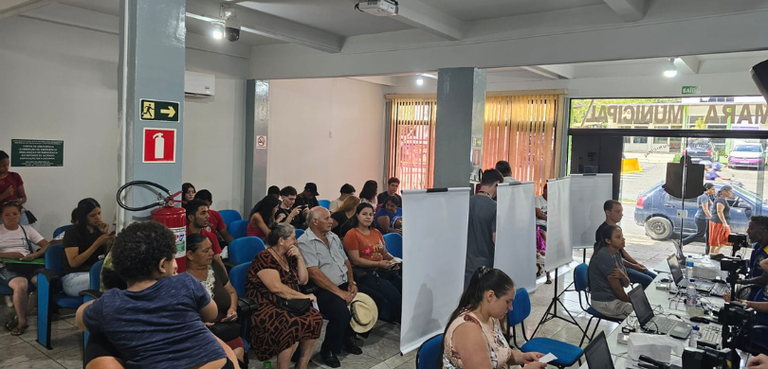 Fotografia de diversas pessoas, entre homens e mulheres, sentadas em cadeiras azuis aguardando a...
