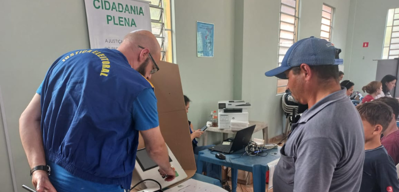 Fotografia em que dois homens aparecem de costas. Um deles, que veste um colete da Justiça Eleit...