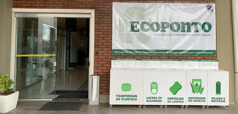 Ao lado de uma porta de vidro, observa-se a estação que serve de depósito para tampinhas de plás...