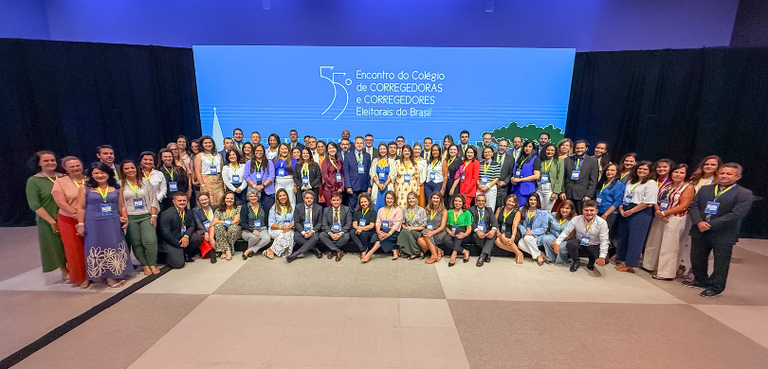 Fotografia de diversas pessoas posando sorrindo em um palco, algumas aparecem em pé e outras sen...